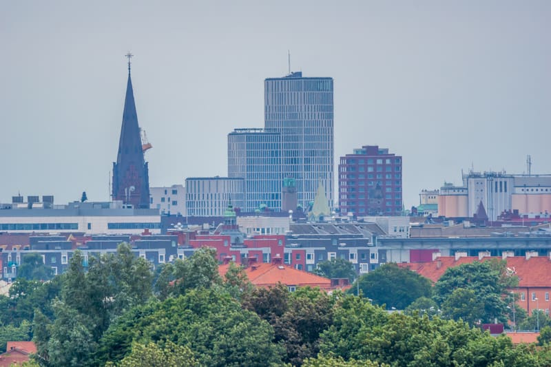 En översiktsbild av en stad med flera byggnader av olika höjd och arkitektur. I förgrunden syns trädkronor med gröna löv och i bakgrunden skymtar en kyrkspira och moderna höghus. Himlen är ljusgrå och en del av byggnaderna har röda tak.