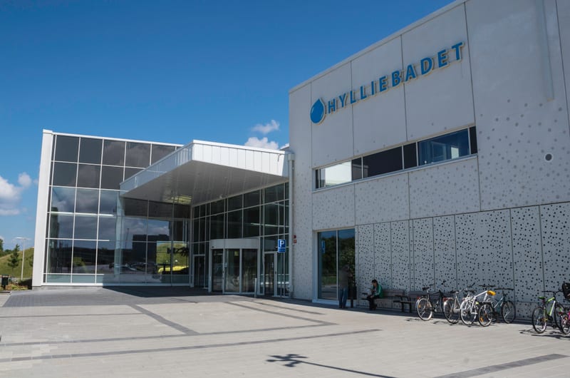 En modern byggnad med stora fönster och en entré med en utskjutande baldakin framför. På fasaden syns texten "HYLLIEBADET" samt en logotyp med en vattendroppe. Framför byggnaden finns ett stenlagt torg med cyklar parkerade och en bänk där två persone