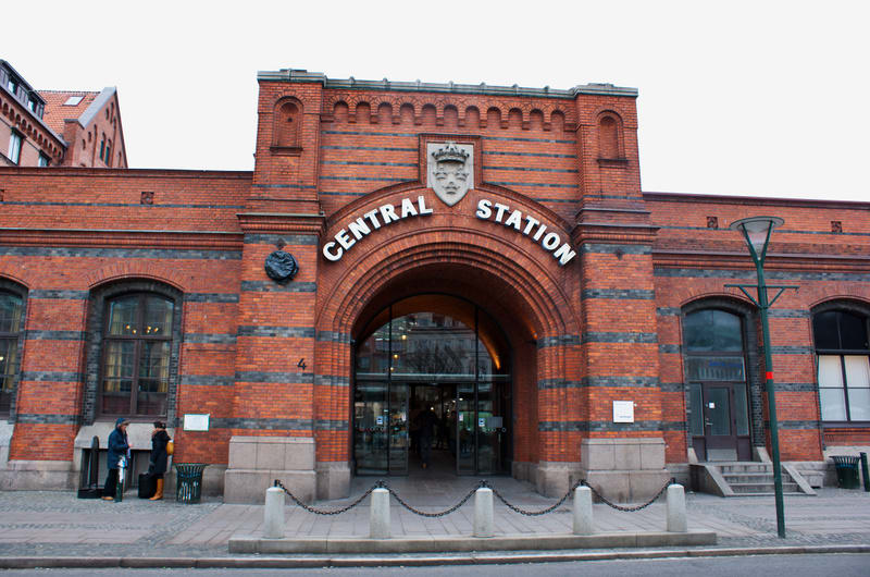 En fasad med rött tegel och ljusa fogar. En stor dörröppning i mitten med bågformade "CENTRAL STATION"-skyltar ovanför. Framför syns två personer, en papperskorg, en lyktstolpe och kedjor mellan betongpelare.