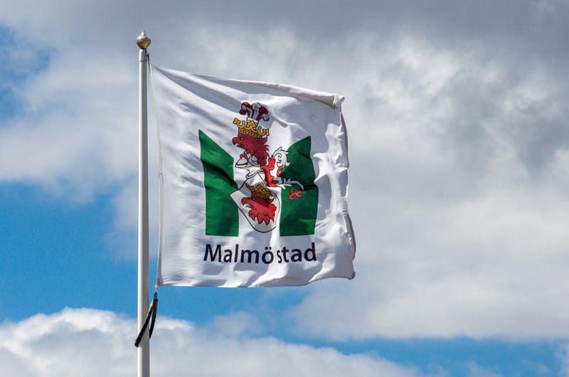 En vit flagga med Malmö stads vapen och texten "Malmö stad" i svart. Vapnet består av ett rött griphuvud med krona samt gröna bokstäver som formar ett 'M'. Flaggan är fäst vid en vit flaggstång och vajar mot en ljusblå himmel med vita moln.