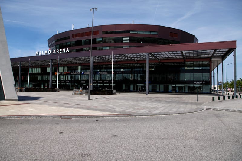 En stor, mörk byggnad med texten "MALMÖ ARENA" på fasaden under en klarblå himmel. Framför byggnaden finns ett öppet torg med stenbeläggning och en asfalterad väg i förgrunden.