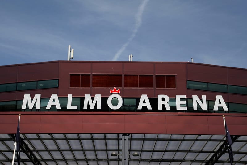 En bild som visar en del av en byggnad med texten "MALMÖ ARENA" i ljusa, stora bokstäver. Ovanför "Ö" syns en röd krona. Byggnaden är rödbrun med fönster och en del av ett takskyltfäste är synligt. Himmelen är blå med vita moln.