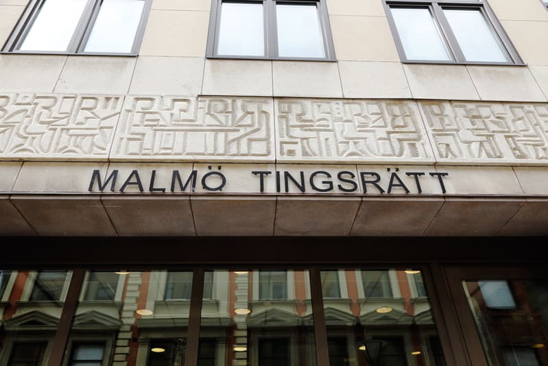En fasad på en byggnad med texten "MALMÖ TINGSRÄTT" i stora svarta bokstäver. Ovanför texten finns ett mönstrat relief och överst syns fönster. Nedanför texten reflekteras en annan byggnad i byggnadens fönster.