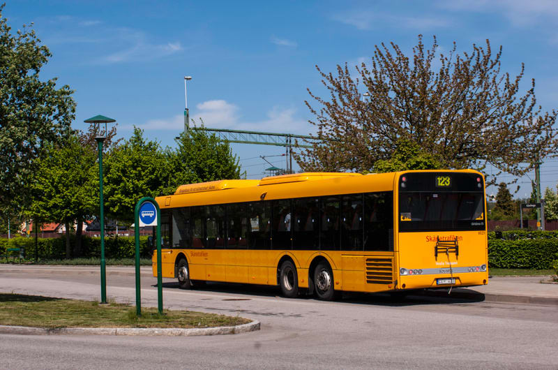 En gul buss från Skånetrafiken står parkerad vid en busshållplats under en solig dag. Busshållplatsen har en skylt och omges av grönska, med träd i bakgrunden. Ett järnvägsspår syns längre bort.