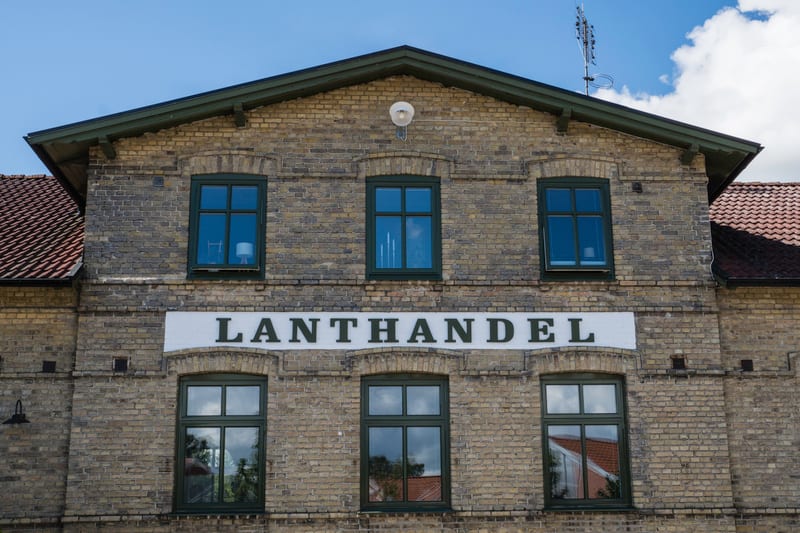 En fasadvy av en tegelbyggnad med gula tegelstenar och gröna fönsterramar. På fasaden står ordet "LANTHANDEL" skrivet i stora, mörka bokstäver på ett vitt band. Taket består av röda takpannor. En TV-antenn syns på taket till höger.