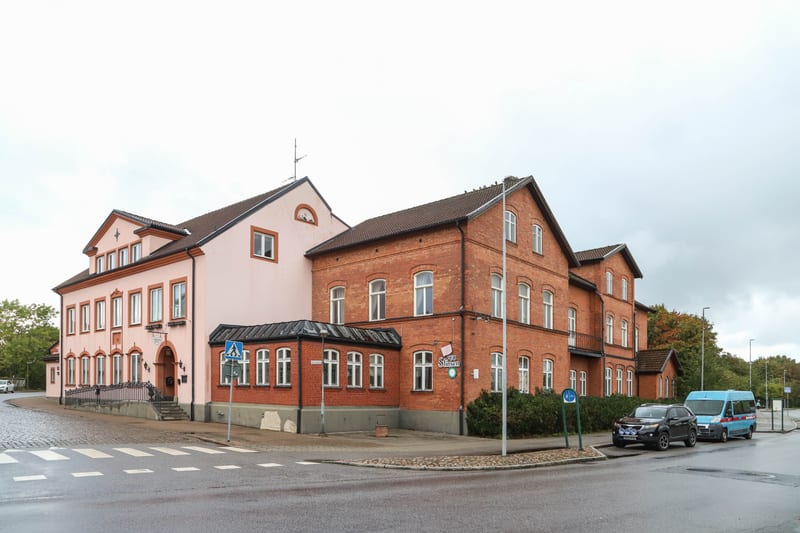 En bild som visar en byggnad med en rosa fasad och en tegelfasad, belägen vid en korsning. Två fordon, en svart SUV och en blå minibuss, står parkerade längs gatan. Vägen i förgrunden har zebrafärger markerade. Växtlighet och en skylt syns också.
