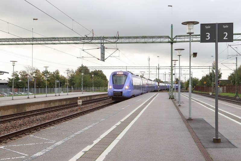 En blått tåg ankommer till en tågstation med flera spår och plattformar under en mulen sky. I förgrunden syns en plattform med lampor och skyltar som indikerar "Spår 3" och "Spår 2". Träd och en byggnad syns i bakgrunden.