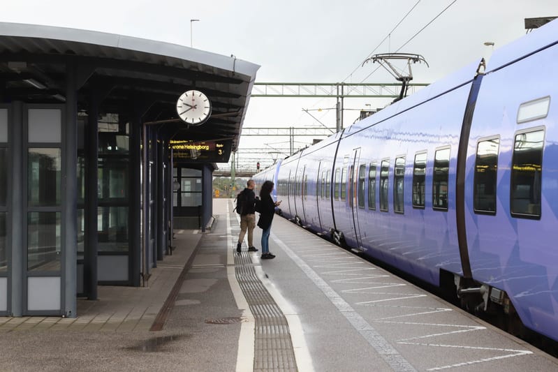 En bild av en tågperrong med ett lila tåg parkerat. Två personer står på perrongen, en man och en kvinna, bredvid tåget. En stationsklocka och en digital skylt som visar 'Helsingborg' och 'Spår 3' syns under ett tak.