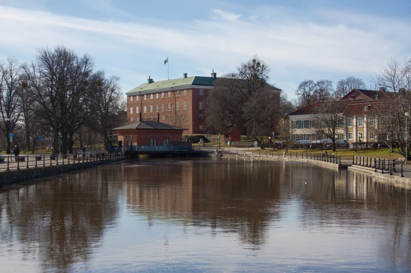 En bild som visar en flod eller kanal med byggnader längs stranden. Till vänster syns ett stort tegelhus med en svensk flagga på taket. Mellan vattnet och det stora tegelhuset finns en mindre tegelbyggnad. Till höger finns ytterligare byggnader och t