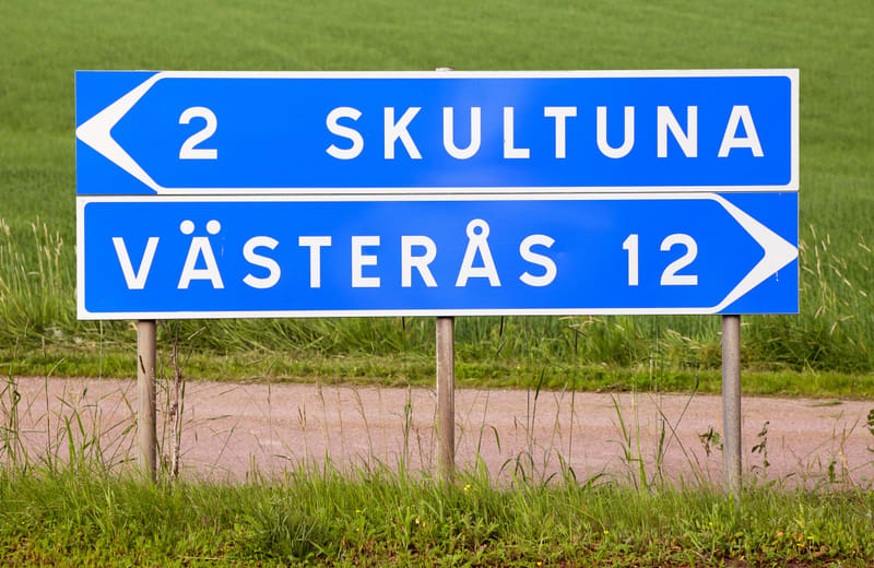 En blå vägskylt med vit text står i gräs bredvid en grusväg. Skylten visar "SKULTUNA 2" med en vänsterpil och "VÄSTERÅS 12" med en högerpil. I bakgrunden syns ett grönt fält.