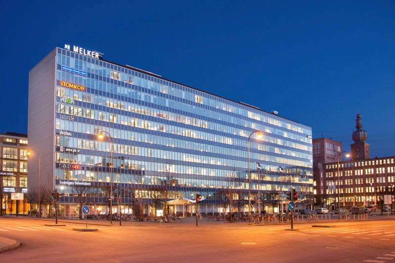 En bild som visar en stor, modern byggnad med många upplysta fönster vid skymning eller natt. Flera företagslogotyper syns på byggnadens fasad, inklusive "MELKER" och "Sveriges Television". Ett torg eller en bred gata med parkerade cyklar och gatlykt