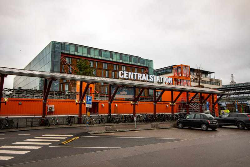 En fasadskylt med texten "CENTRALSTATION" syns framför en byggnadskonstruktion som består av orangea containrar och en grått tak. Bakom containrarna syns en större, modern byggnad med glasfasad och träpaneler. Framför containrarna står flera cyklar p