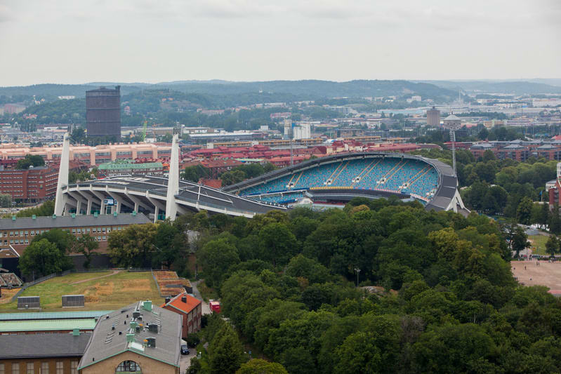 En översiktsbild av en stad som visar en stor idrottsarena med blå sittplatser, omgiven av grönskande träd. I bakgrunden syns fler byggnader och kuperad terräng under en molnig himmel. Ett stort brunt gasverk kan även ses i bildens vänstra del.