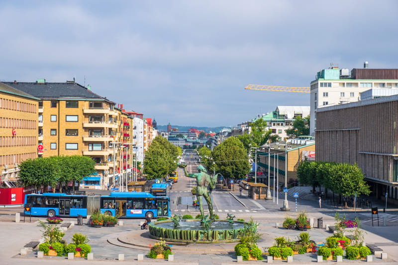 En översiktsbild av en stadsgata med en staty i förgrunden. Blå bussar, flerbostadshus och träd kantar gatan under en himmel med viss molnighet. En byggkran syns i bakgrunden.