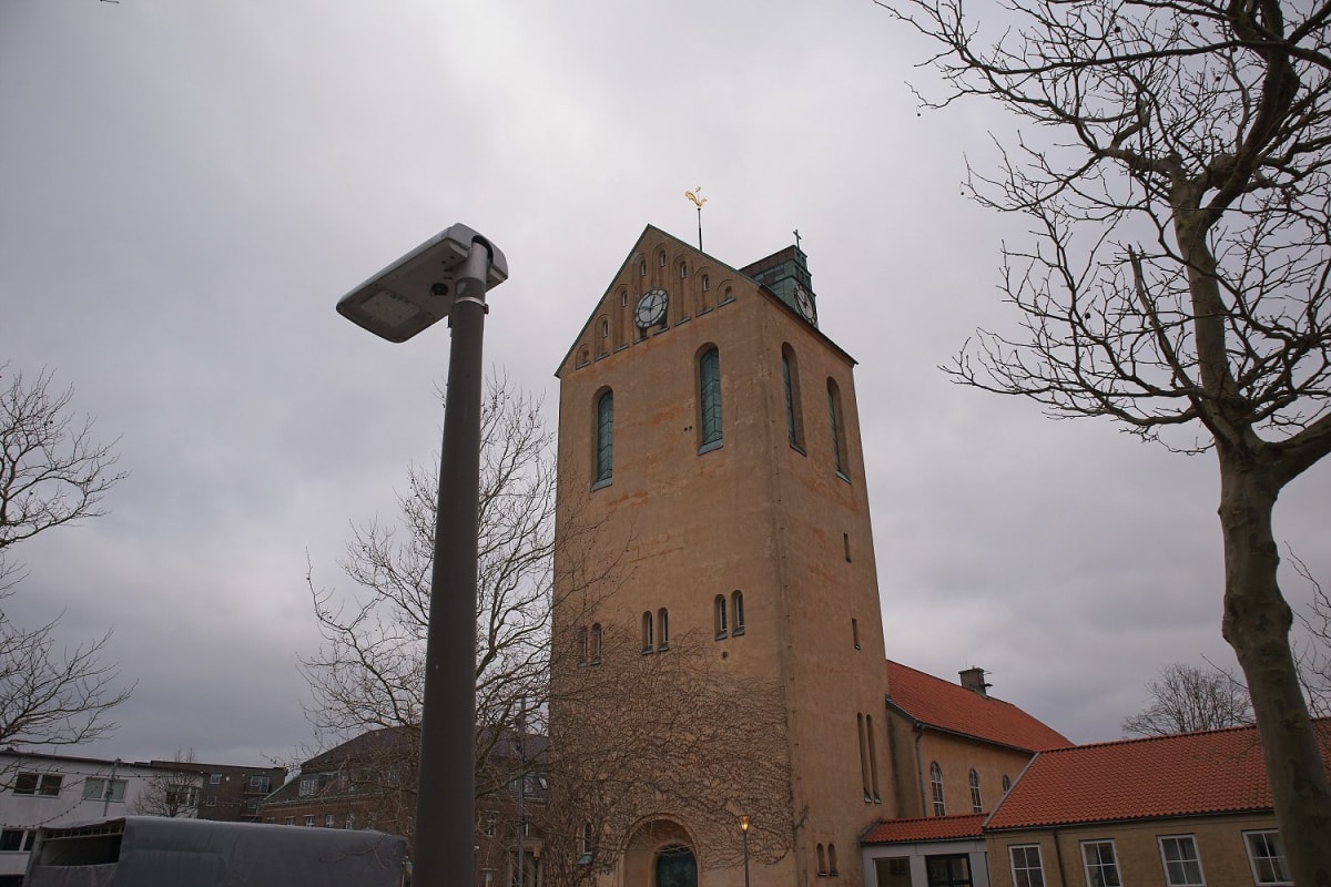 En stor, sandfärgad kyrkobyggnad med ett högt torn med urtavlor syns under en gråmulen himmel. Nakna trädgrenar och en gatlykta ramar in byggnaden. En del av kyrkans tak syns baktill, täckt med tegelpannor och med en utbyggnad till höger.