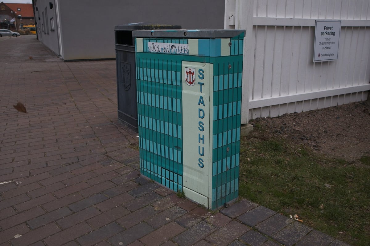 En blågrön elbox målad som ett hus med texten "STADSHUS" och ett ankarsymbol syns vid en tegelbelagd gångväg. I bakgrunden finns en mörk soptunna, en vit staketvägg med en skylt för "Privat parkering" och en gräsplätt med barr och löv. En ljusbrun by