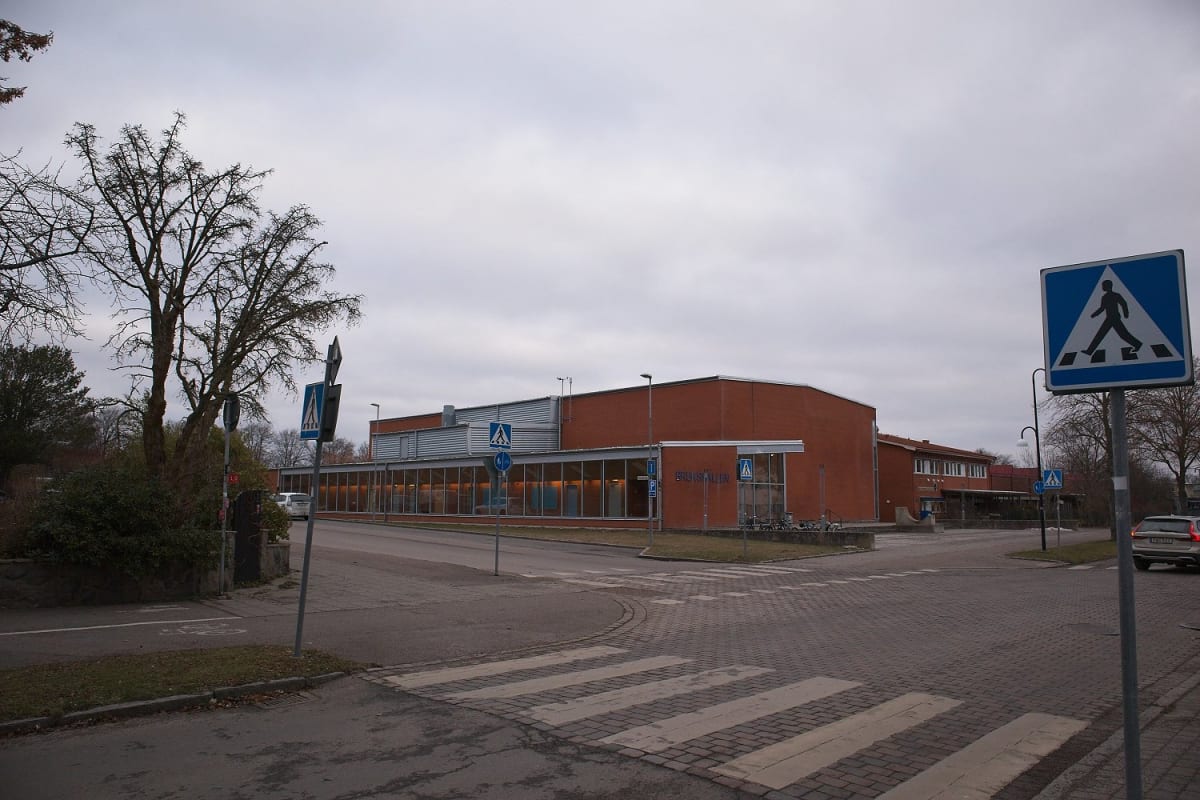 En utomhusbild som visar en gatukorsning med övergångsställen och skyltar. En stor tegelbyggnad syns i bakgrunden, med texten "BRUKSHALLEN" på fasaden. Till vänster om byggnaden finns träd utan löv och en buskage.