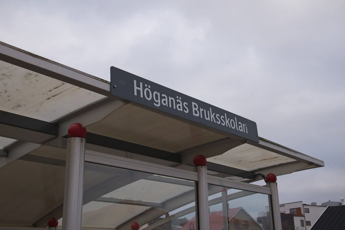 En bild tagen underifrån visar en busshållplats med en skylt där det står "Höganäs Bruksskolan" med vit text på en mörkgrå bakgrund. Taket på busshållplatsen är transparent med metallramar. I bakgrunden syns en gråmulen himmel och delar av byggnader.