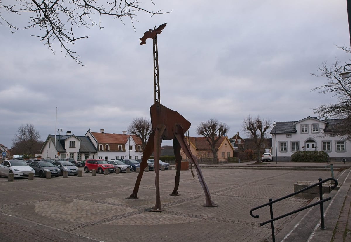 En stor, rostfärgad skulptur föreställande en giraff står på ett torg framför parkerade bilar och hus. Träd utan löv och en gråmulen himmel syns också.
