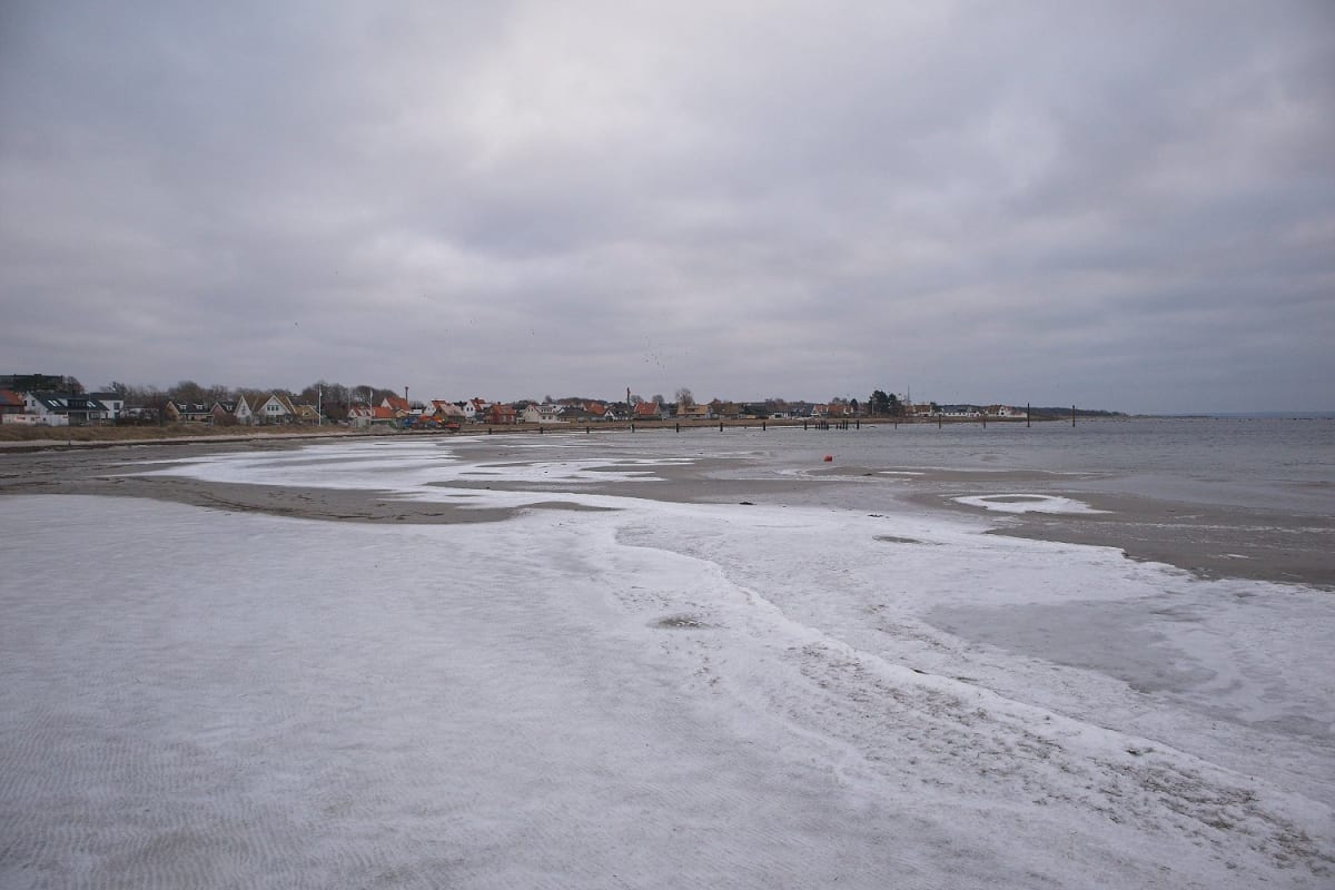 En bild som visar en snötäckt strand med ett vattendrag till höger. I bakgrunden syns hus och byggnader längs vad som kan vara en kustlinje, under en grå och molnig himmel.