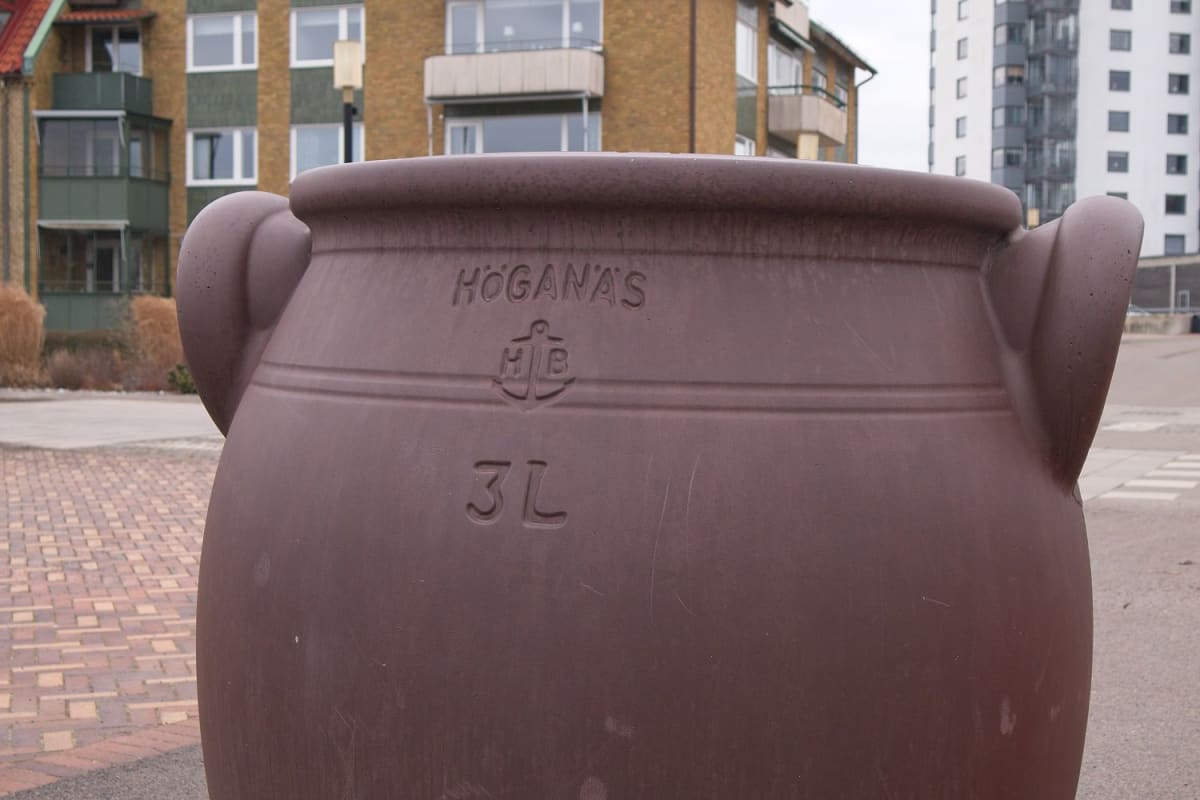 En stor, brun kruka med handtag på sidorna står i förgrunden, med texten "HÖGANÄS" och "3L" samt en symbol med ett ankare. I bakgrunden syns bostadshus med fasader i tegel och vita fönsterkarmar, samt grå himmel.
