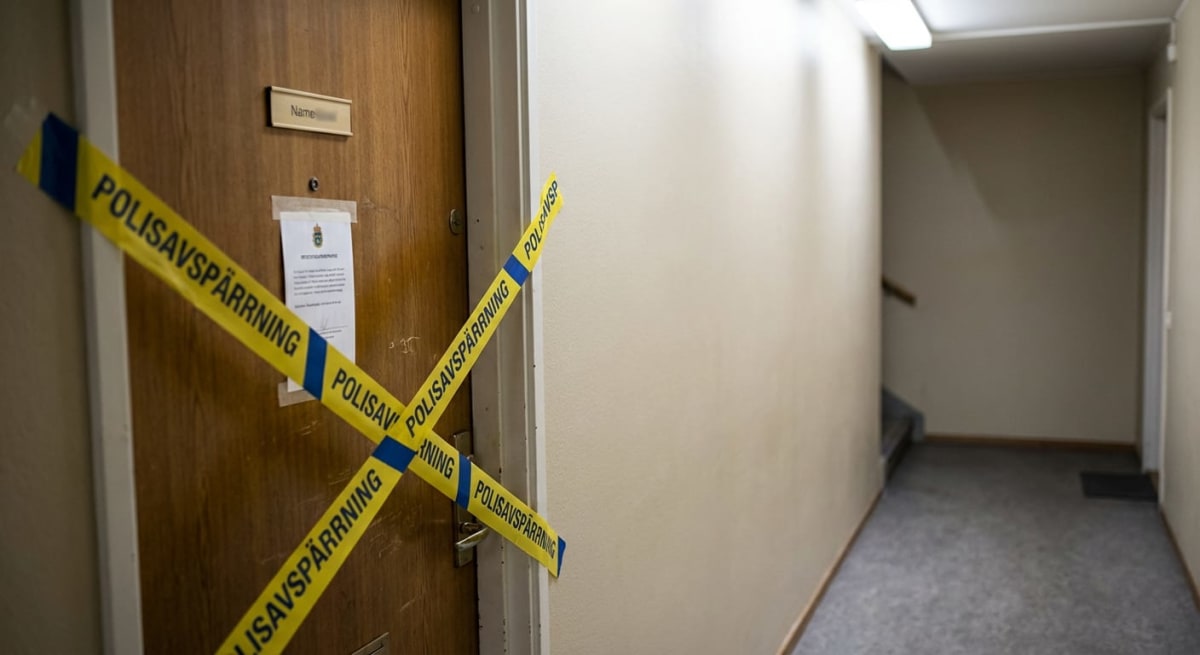 En gul, blårandig "Polisavspärrning"-tejp sitter tejpad i ett kors över en ljusbrun trädörr i en inomhuskorridor. Ett papper med en kunglig krona och text är tejpat på dörren. Ljus vägg till höger och en matta på golvet. En namnskylt är synlig ovanfö