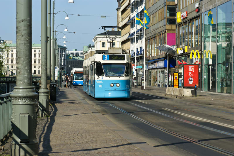 En ljusblå spårvagn med nummer 319 och skylten "3 Kålltorp" står på spår i en stadsmiljö. I bakgrunden syns byggnader, en buss, människor på trottoaren och svenska flaggor. En McDonalds-skylt är synlig till höger.