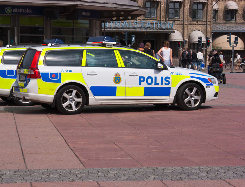 Två svenska polisfordon, vita med blå och gula markeringar, står parkerade på en stenlagd yta. I bakgrunden syns en byggnad med skylten "RALSTATION" och trafikanter som rör sig i området. Hjul och däck på fordonen syns tydligt.