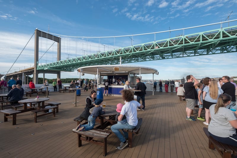 En utomhusscen under dagen visar en grupp människor som befinner sig på en byggnad eller anläggning med trägolv och ett utomhuscafé/bar, med en stor grön bro i bakgrunden. Flera personer sitter vid träbord och bänkar, medan ett antal står vid baren,