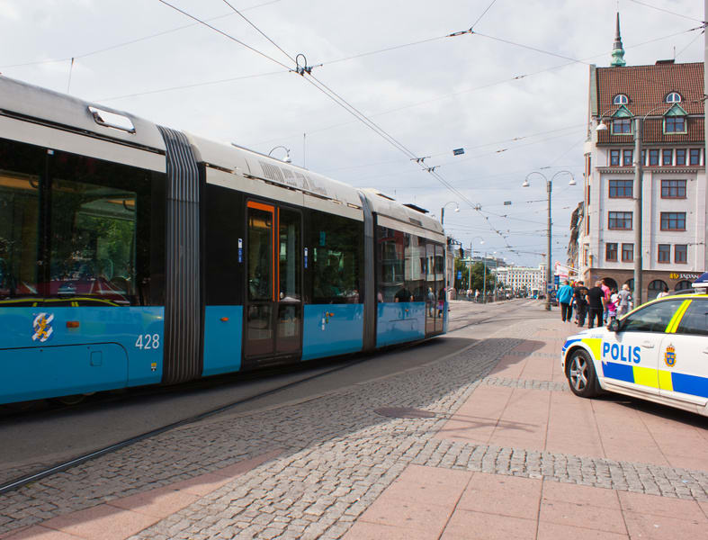 En gatuscen där en blå och vit spårvagn kör på räls till vänster i bilden, medan en vit och gul polisbil står parkerad till höger. I bakgrunden syns en byggnad i äldre arkitekturstil samt flera personer som går på trottoaren. Över gatan hänger elkabl