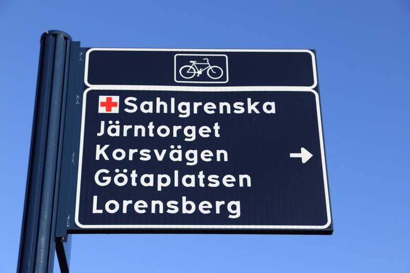 En blå skylt med vit text som visar vägen till Sahlgrenska, Järntorget, Korsvägen, Götaplatsen och Lorensberg. En röd symbol för sjukhus och en cykelsymbol syns också på skylten. Skyltstolpen är synlig till vänster, och en ljusblå himmel utgör bakgru