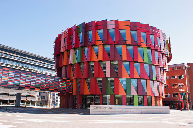 En modern byggnad med en fasad av färgglada, triangulära paneler i rött, orange och grönt under en klarblå himmel. Entrén har en skylt med texten ”CHALMERS”. Till vänster syns delar av en annan byggnad med en horisontell gångbro.