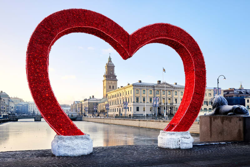 En stor, röd, glittrig hjärtformad installation står i förgrunden, delvis skymmande en stadsmiljö med en kanal eller flod i mitten, kantad av byggnader. En klocka syns på en av byggnaderna i bakgrunden. En statyett av ett liggande lejon syns till hög
