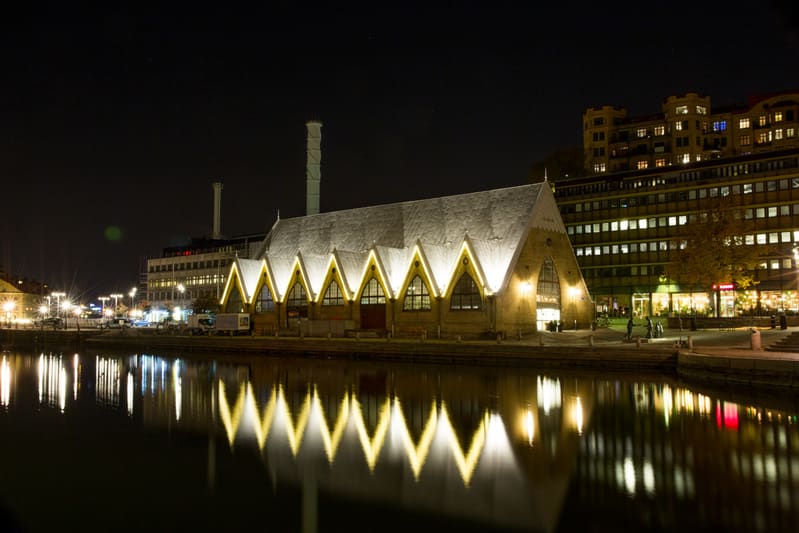 En nattbild som visar Feskekôrka, en fiskmarknadsbyggnad, vid en kajkant. Byggnaden är upplyst med ett gult sken som reflekteras i vattnet framför den. I bakgrunden syns fler byggnader med upplysta fönster samt två skorstenar. En del av himlen syns h
