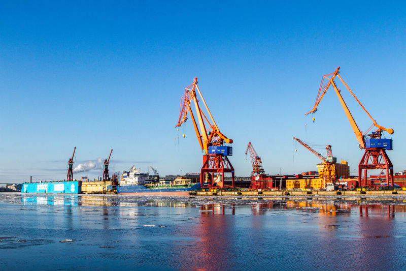 En bild av en hamn med flera stora lyftkranar i orange och rött samt en blå containerbåt förtöjd vid kajen. Vattenytan är delvis täckt av isflak och himlen är klarblå.