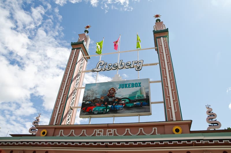 En bild som visar en stor portingång med texten "Liseberg" överst. Nedanför syns en stor reklamskylt med ordet "JUKEBOX" och bilder på klassiska bilar. Två höga torn med dekorationer flankerar skylten. Flaggor i rosa och grönt vajar ovanför texten. L