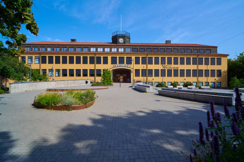 En gul tegelbyggnad med rött tak och en klocka i mitten, under en klarblå himmel. Framför byggnaden finns ett torg med stenläggning, planteringar och sittbänkar. Skyltar på byggnaden indikerar "Växthuset" och "Västerås Kulturskola".