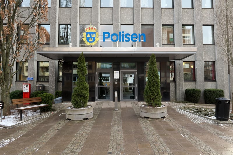 En fasadbild av en byggnad med texten "Polisen" och en gul polissymbol ovanför entrén. Framför entrén finns två avlånga gröna buskar i grå krukor, en träbänk, en svart soptunna och lite snö på marken. Fasadens yta är grå och har flera fönster i rader