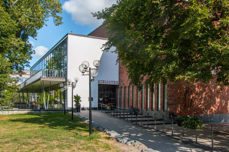 En fasad på en byggnad med texten "BIBLIOTEK" ovanför en entré, med stora fönsterpartier, balkong, gräsyta, träd och lampor i förgrunden.