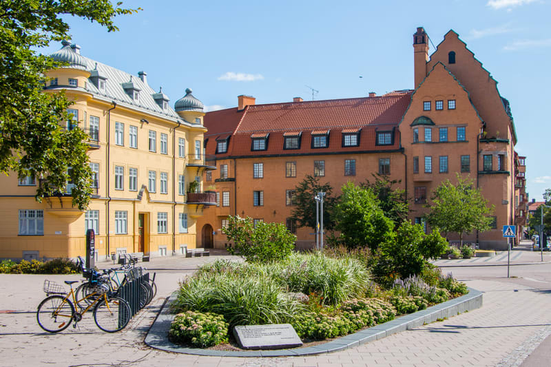 En ganska solig eftermiddagsbild som visar en gatukorsning och ett inhägnat område med grönska i ett bostadsområde. Flerfamiljshus i olika färger; närmast till vänster i gult, medan husen i bakgrunden är tegelröda och tegelorangea. Cyklar står parker