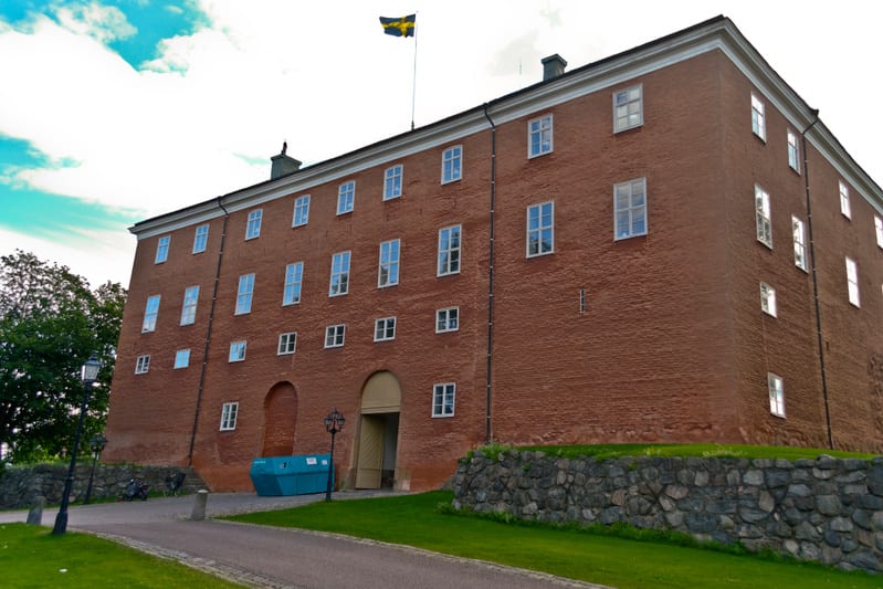 En stor röd tegelbyggnad med många fönster syns under en ljusblå himmel med moln. En svensk flagga vajar från taket. Framför byggnaden finns en gräsklädd sluttning, en stenmur, en grusgång och en turkos container. Ett par lyktstolpar och träd syns oc