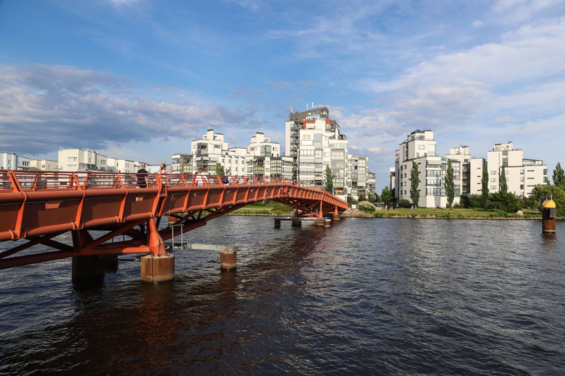 En röd bro sträcker sig över ett mörkt vatten under en klarblå himmel med moln. Längs flodkanten, på andra sidan bron, står modern arkitektur med vita byggnader. En gul boj syns i vattnet till höger om bron. Flera personer står på bron.