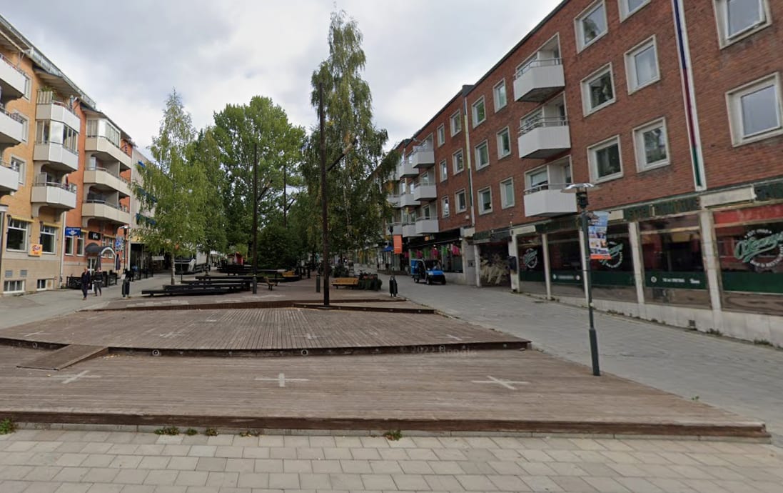 En gatuvy som visar en bred gata med byggnader på båda sidor. I förgrunden finns en trädäckad yta med sittmöbler. På den högra sidan syns en röd tegelfasader med butiker på markplan och lägenheter ovanför. På den vänstra sidan syns en ljusare byggnad