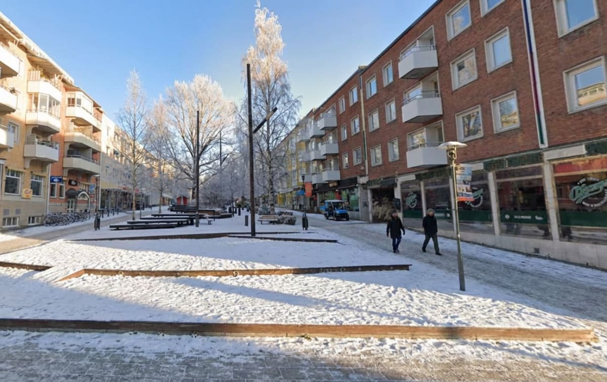 En gata täckt av snö med byggnader på båda sidor och träd med frostigt lövverk, belyst av en klarblå himmel. Två personer går på trottoaren till höger, och cyklar står parkerade på vänster sida. En blå mindre lastbil syns längre bort på gatan.