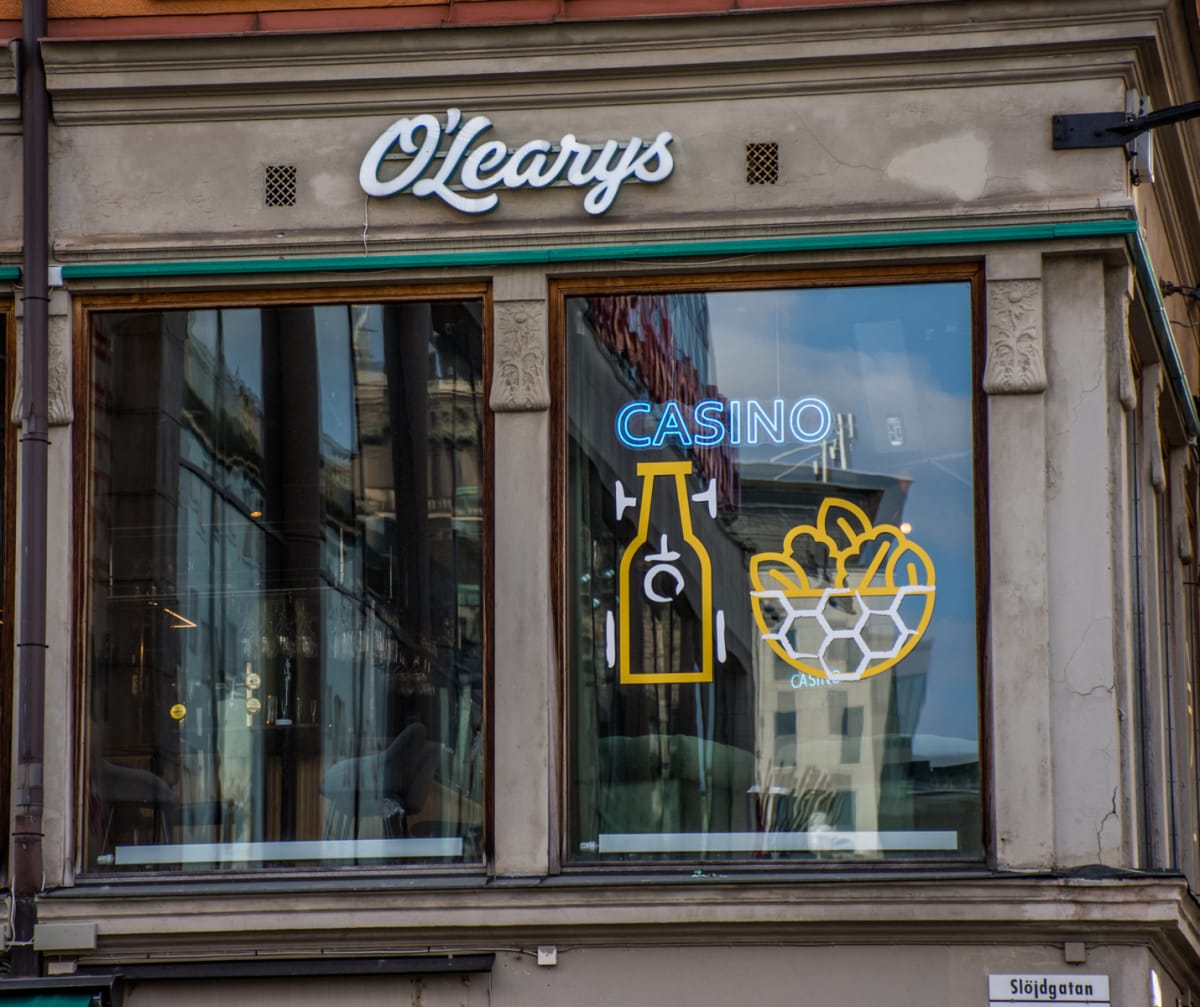 En fasadvy av en byggnad med skylten "O'Learys" ovanför två stora fönster. I det högra fönstret syns en neonlampa med texten "CASINO" och symboler föreställande en flaska och en skål med mat. En gatuskylt med texten "Slöjdgatan" är synlig längst ner