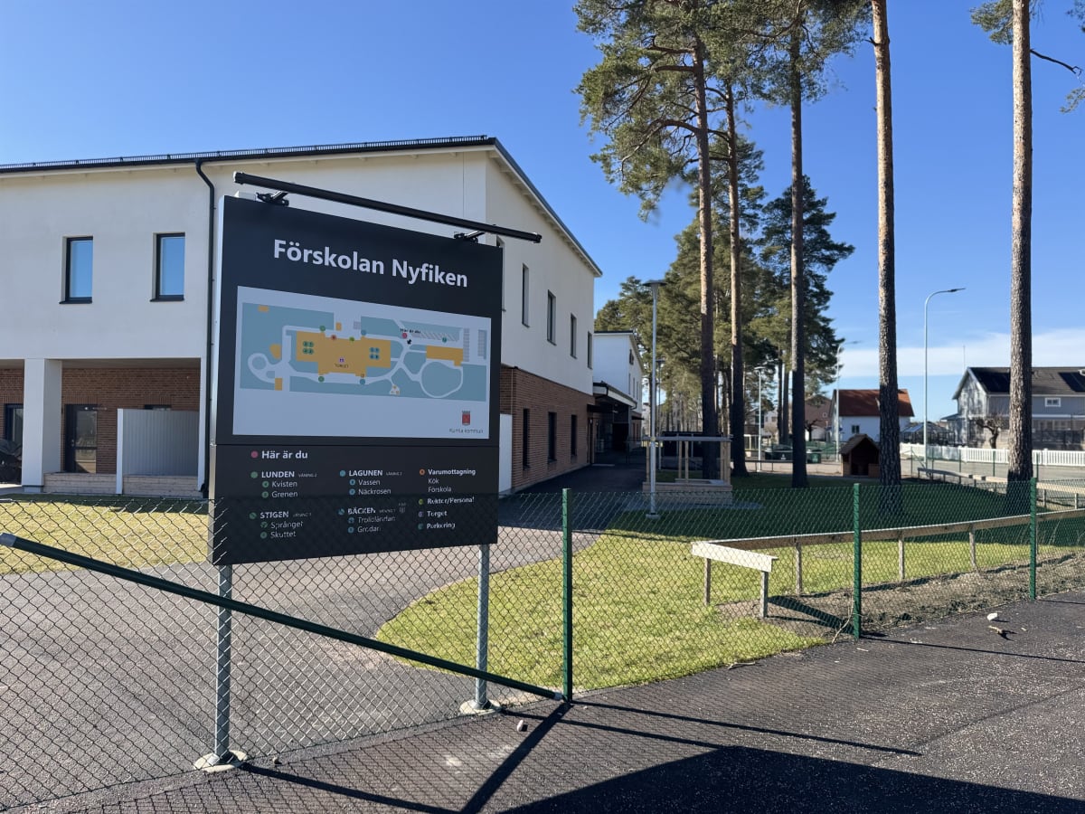 En utomhusbild tagen en solig dag som visar upp en stor svart skylt för "Förskolan Nyfiken" med en karta. Bakom skylten finns en byggnad med vita väggar och svarta fönster, samt höga tallar. Ett gräsbevuxet område avgränsas av ett grön maskstängsel i