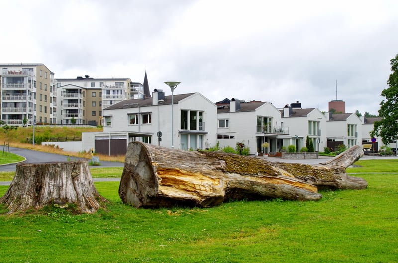 En stor, fälld trädstam ligger på en gräsmatta i förgrunden, bredvid en trädstubbe. I bakgrunden syns moderna bostadshus, både flerfamiljshus och radhus, under en gråmulen himmel. En väg slingrar sig genom området.
