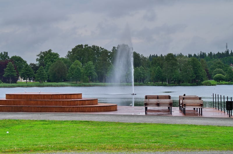 En utomhusbild som visar en sjö med en fontän i mitten, omgiven av grönskande träd och en gråmulen himmel. I förgrunden finns en träterrass med trappsteg, bänkar, en brygga och ett gräsområde.