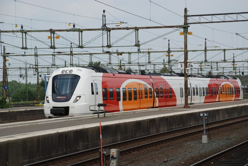En modern designad tåg som står vid en perrong. Tåget har röda och orangea detaljer på en vit bakgrund, med fönster längs sidorna. Ovanför tåget syns ett komplext nät av kontaktledningar och stolpar, och i bakgrunden skymtar trädgrupper. På plattform