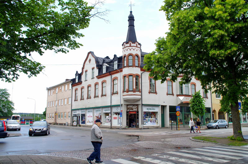 Bilden visar gatukorsningen framför en större vit byggnad med ett torn och flera butiker på bottenplan. En person går över ett övergångsställe i förgrunden. I bakgrunden syns bilar och träd med gröna löv.